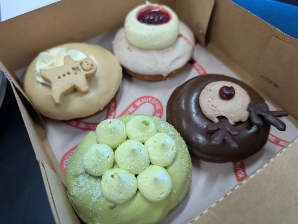Photo of 4 donuts in a brown cardboard box, taken from the corner at a 45° elevation. The box is diagonally oriented making one of the donuts look big and delicious, like an offering to you. They are placed on top of a pink printed greaseproof sheet with a pattern of ribbons with writing. The writing says "you're beautiful". The donuts are all Christmas themed. Clockwise from the front: pastel green icing with matching buttercream "kisses" piped in a ring of six with a centre one. I guess it should be a snowflake. The next one has a soft latte brown glaze with a plain white whipped cream rosette that a tiny gingerbread man is placed on, off-centre. The third one has a speckled pink white chocolate dip with a large buttercream blob piped on top, about half as wide as the donut. It has an indent that is filled with red jam. The final donut has dark chocolate dip with a light brown buttercream blob just covering the hole. Two chocolate reindeer "antlers" have been stuck in at the top and a small blob of red jam serves as a nose.