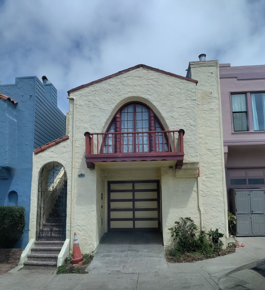 A 2 level home on a busy street in San Francisco. The narrow driveway leads directly to a recessed garage door at the center of the home. On the left an exterior staircase leads to the 2nd floor. There is an archway about 4 steps up, with a beautiful wrought iron security screen. The 2nd floor of the facade features a small balcony with an oblong archway lined with glass paned doors and windows. The front of the home is a pleasant yellowish cream color with a sort of rusty burgundy colored trim on the garage, balcony fence, and trim on the balcony. There is an orange traffic/safety cone between the opening that leads to the garage and the exterior staircase, and some green plants are growing in front of the house to our right of the garage recess. The blue sky above is obscured by fluffy white clouds. We can also see part of a dusty rose colored home on our right, and a dark sky blue home on our left 