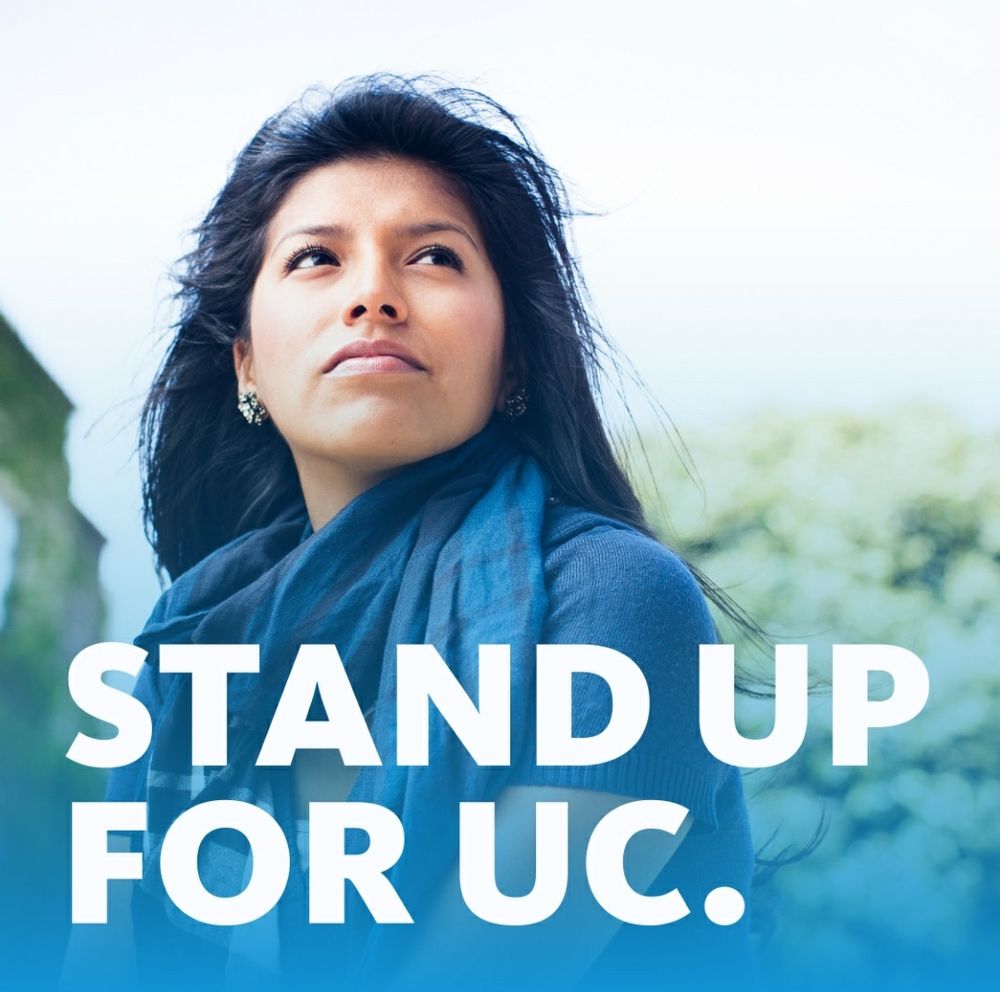 Graphic shows a woman with long, dark windswept hair facing us, eyes looking up and to our right. She is wearing blue and appears to be standing in front of some blurred green bushes. There is bold white text overlayed at bottom, reading STAND UP FOR UC.