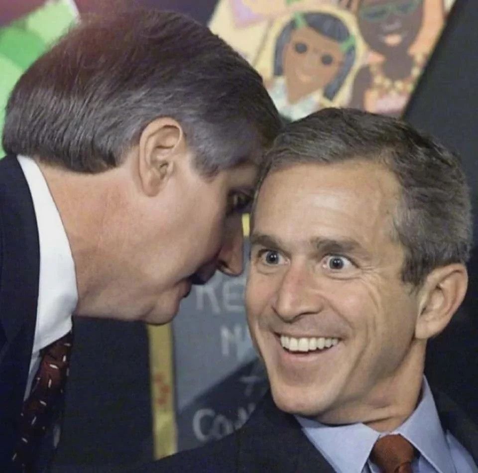Dois homens brancos, sendo um deles George W. Bush. Ele está sentado e ouve do outro homem um recado próximo ao seu ouvido. A foto é editada para que Bush apareça sorrindo e com olhos arregalados.