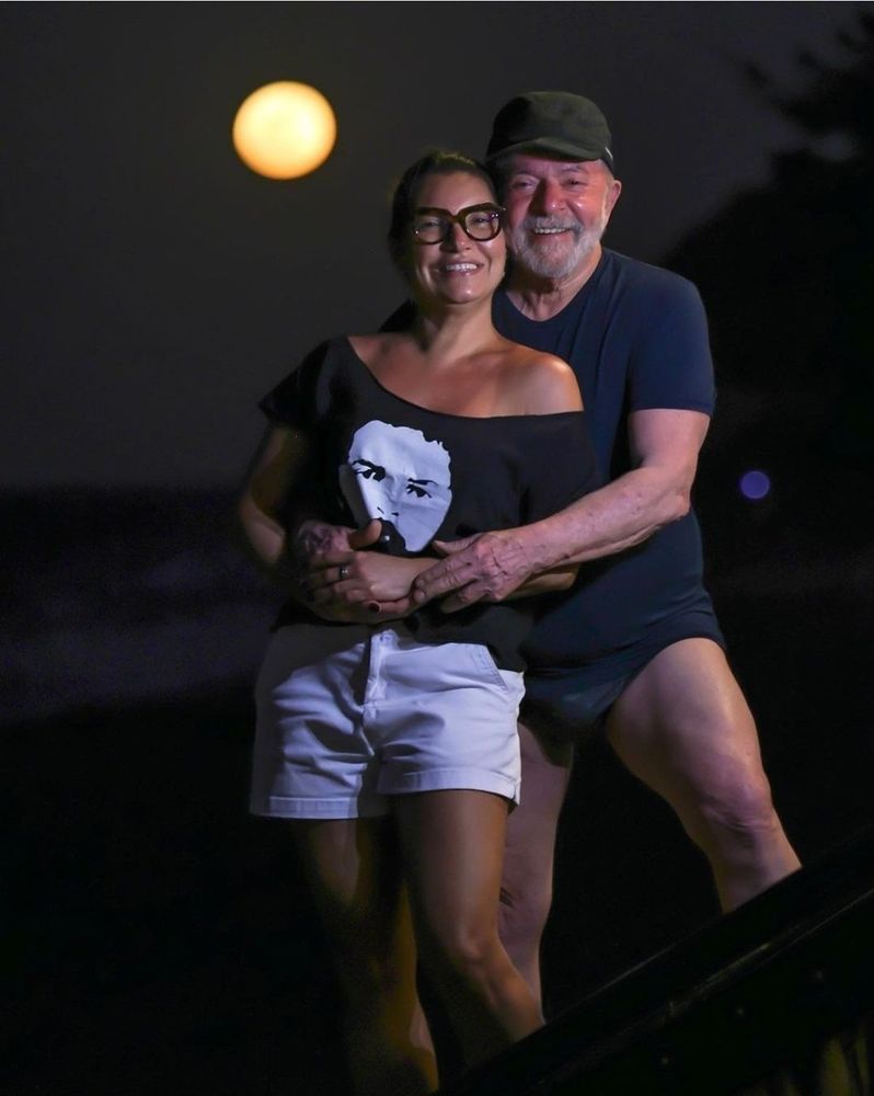 Janja e Lula sorrindo para a foto. Ele a abraça por trás. Ela veste uma camiseta preta com o rosto dele jovem. Ele usa um boné, camiseta e sunga, mostrando as coxas. Ao fundo, o mar de noite e a lua amarela desfocada.