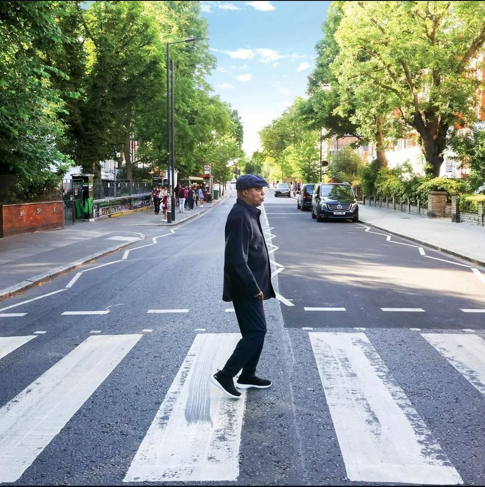 Milton Nascimento atravessando a icônica Abbey Road, visto de perfil do mesmo ângulo que na capa do álbum dos Beatles. Ele é um homem negro, usa roupas pretas, agasalho e uma boina. É uma rua muito arborizada, com alguns carros estacionados e num dia ensolarado.