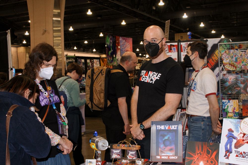 sean at the haunted table booth wearing a black triangle t shirt and mask, glancing at the camera as con goers peruse dice, books, and props from triangle agency. ben is helping a customer as he stands in front of prints in the background 