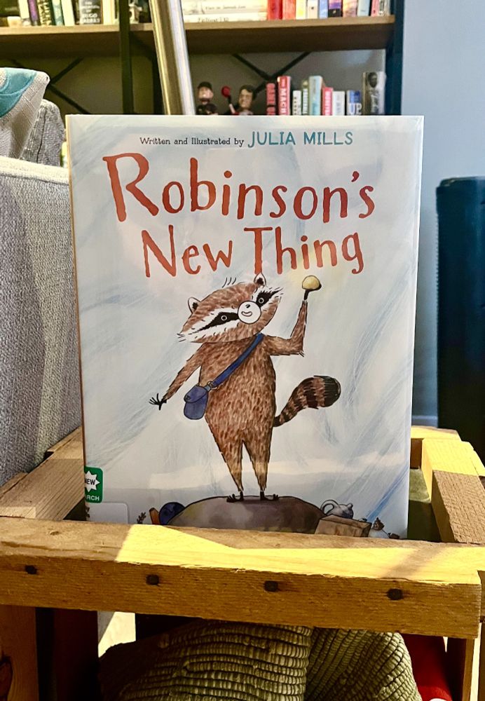 Photo of a picture book. The cover features a brown raccoon standing on top of a rock, a blue bag slung across his torso, proudly holding up a small pebble. At his feet are various knickknacks, plus a snail friend. Text reads, "Written and illustrated by Julia Mills, Robinson's New Thing."