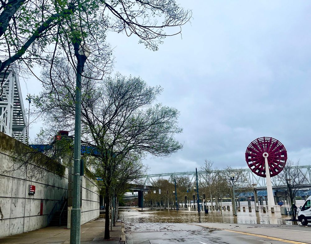 Photo of a flooded street (Mehring Way in downtown Cincinnati) flanked by the edge of a stadium on the left and a riverboat wheel statue on the right. 