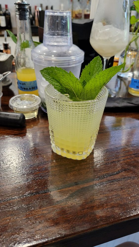 A wooden bar top with a glass tumbler filled with a citrus mocktail and garnished with mint leaves. In the background is various ingredients for making mixed drinks.