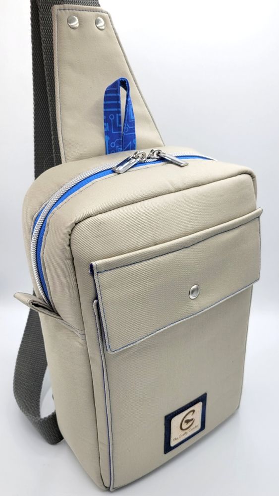 A beige or grey colored bag with a blue zipper that is shaped like a rectangle. It has a front flap pocket and a sling style strap for wearing across the body.