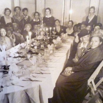 Black and white photo circa 1920 of approximately 25 Black women dressed up for tea in long dresses and hats. They are seated around a long table and many are smiling for the photograph. The dining table is draped in a fine white tablecloth, place settings are out, and many candles are lit.