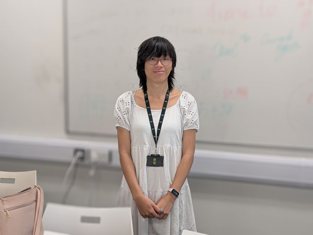 Kristina, in a white dress and wearing an X1 Labs lanyard, standing in front of a whiteboard. 
