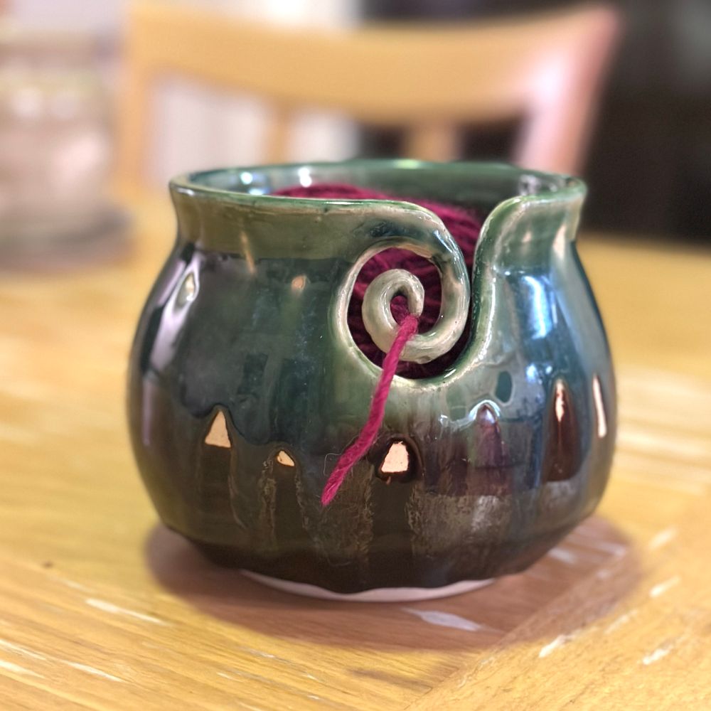 A ceramic bowl to hold yarn for knitting or crocheting. The bowl is has a round belly and a vertical lip with a spiral cut out for capturing the a thread. It is decorated at the top with blue and green glaze which drips down over a dark brown bottom half. A maroon ball of yarn is inside the bowl with a short thread passing through the center of the spiral. 