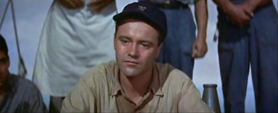 A navy ensign played by jack lemmon squats on his read while wearing a sweat-stained shirt and blue ball cap. 