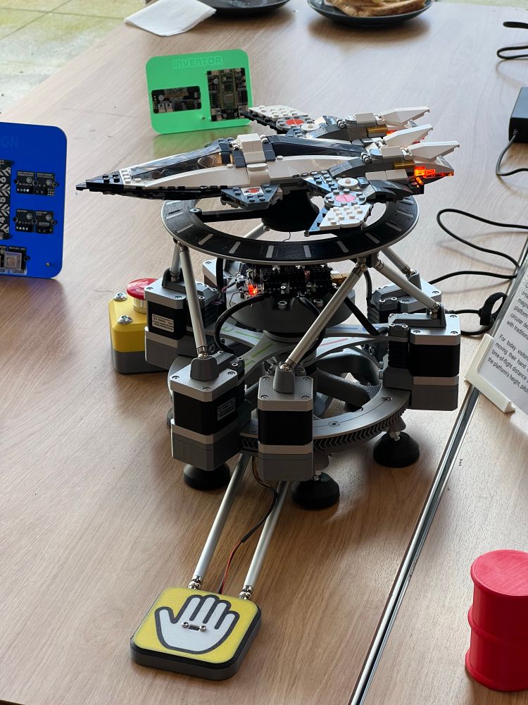 A Lego spaceship mounted on top of an assembly of six stepper motors around a base ring, a six axis motion platform. The engines are lit up.