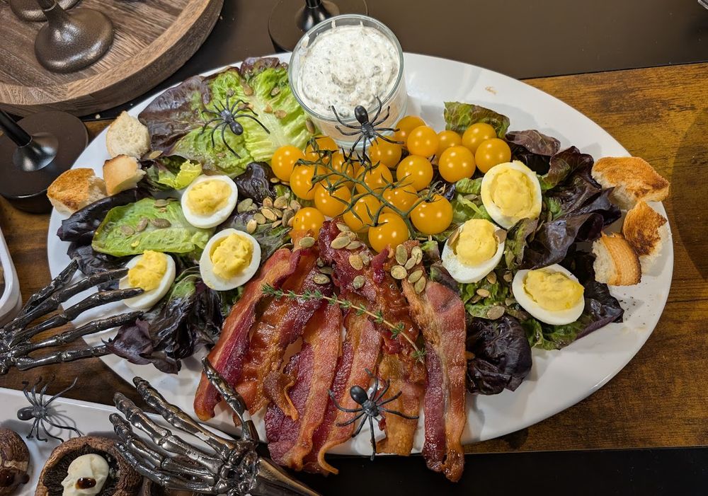 A large white platter features a Halloween-themed salad arranged with sections of leafy greens, halved hard-boiled eggs, yellow cherry tomatoes on the vine, crispy bacon strips, toasted bread pieces, and scattered pumpkin seeds. A glass container of creamy dip sits at the top center of the plate. Several plastic black spiders are placed on the salad as decoration, and a pair of metal skeleton hands rest at the bottom left corner of the platter.
