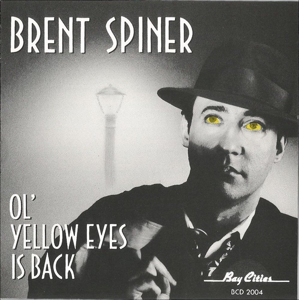Album cover for *Ol' Yellow Eyes Is Back* by Brent Spiner. The image is in black and white, except for Spiner's bright yellow eyes, which stand out. He is dressed in a vintage-style suit and tie with a fedora, holding a cigarette and looking slightly off to the side. A streetlamp glows faintly in the background. The text on the cover reads 