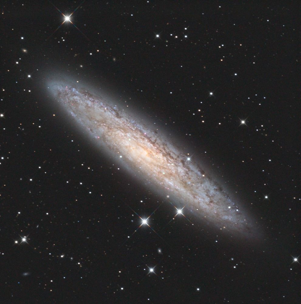 The Sculptor Galaxy, a nearly edge on spiral galaxy in the constellation of the same name. It cuts diagonally across the photo from upper left to lower right, and has blueish outer arms and a golden core. Its surrounded by a sparse star field.
