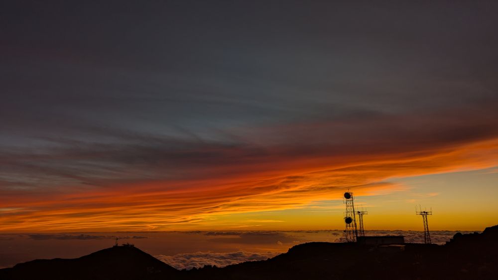 The view west from the Haleakala Summit, grey clouds above and vivid orange and red bands near the horizon. Some antennas and rocky hills in the foreground 