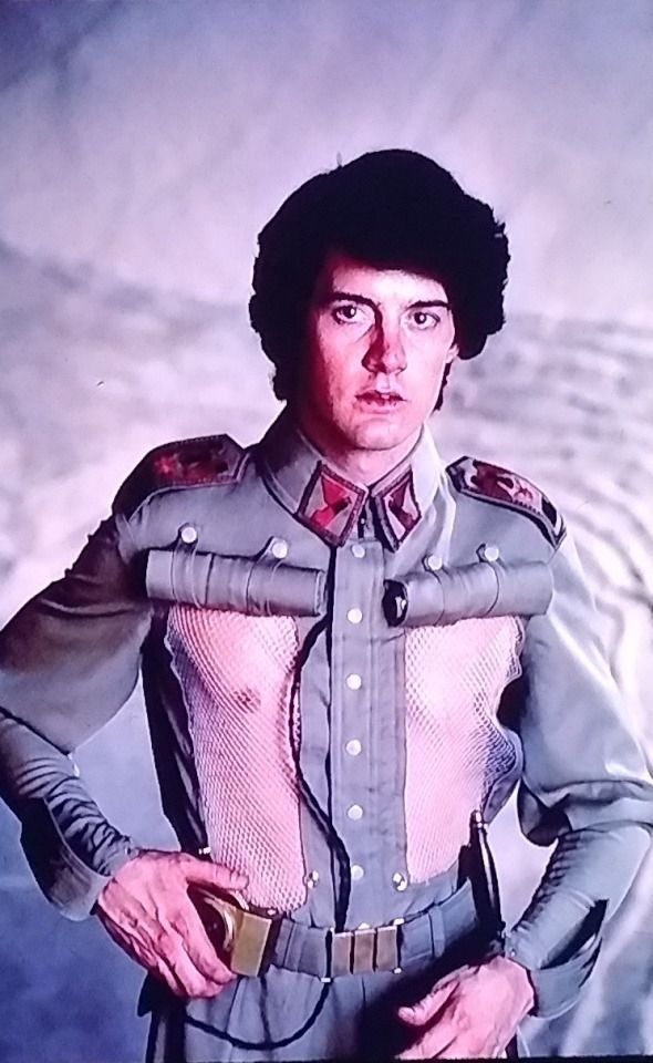 A dark-haired man wearing a military uniform and fiddling with a brass device on his belt. His shirt has two vertical panels that are rolled up to reveal mesh underneath. His nipples can be seen through the mesh. 