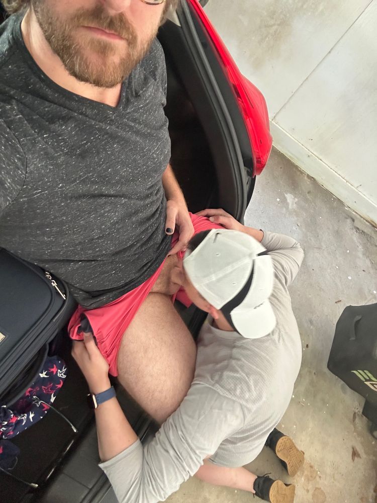 Me on my knees behind a car in a garage while my husband sits on the back of the car with his dick down my throat