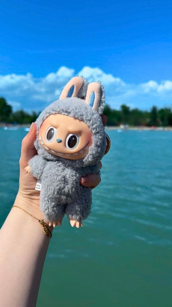 Scrunchies hand holding up her sea salt coconut blue labubu overtop of the lake water looking to beach shoreline with a thick tree line low clouds and blue sky.