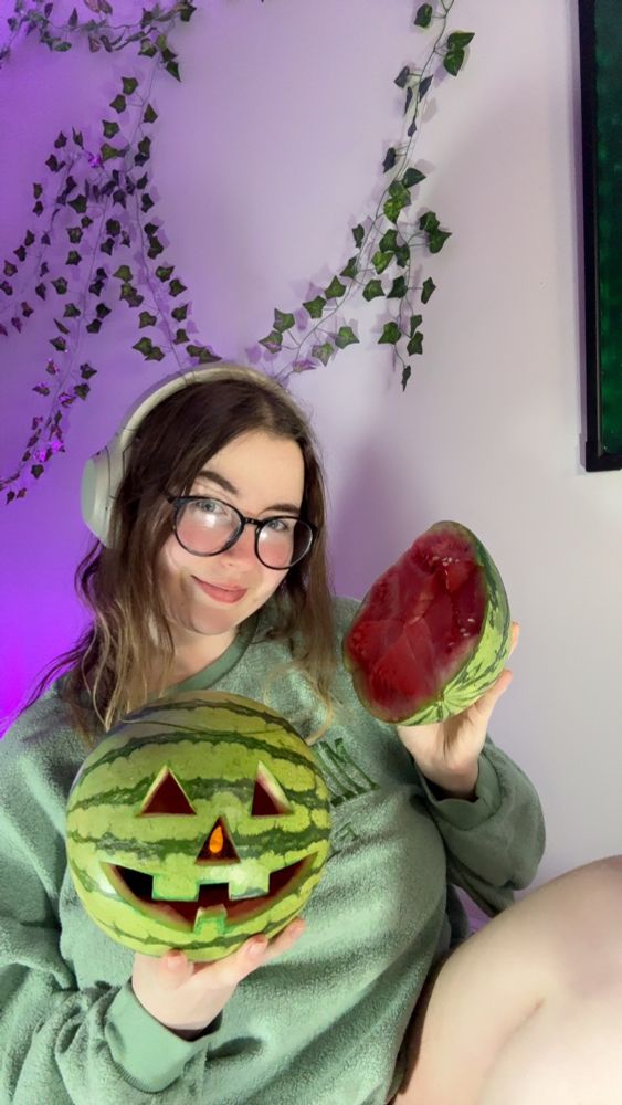 Scrunchie holding a watermelon that’s been carved to look like a jack-o’-lantern i. One hand and holding half of a watermelon that she crushed with her thighs in the other hand 