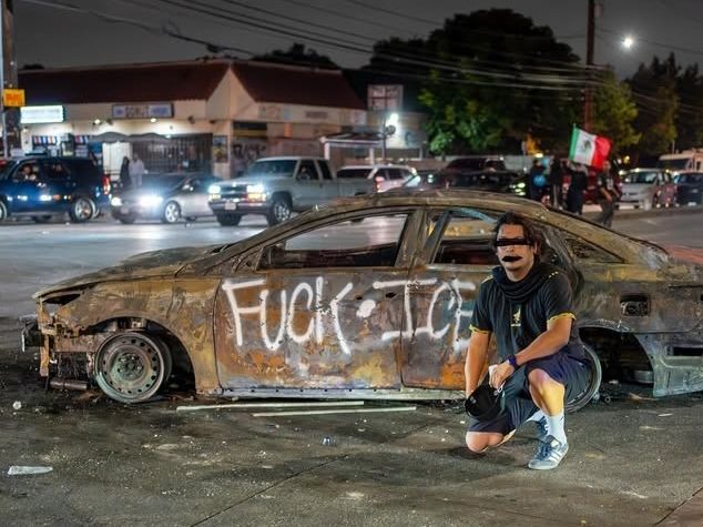 Protestor kneels in front of burnt out car, graffitied with a slogan which reads "FUCK.ICE"