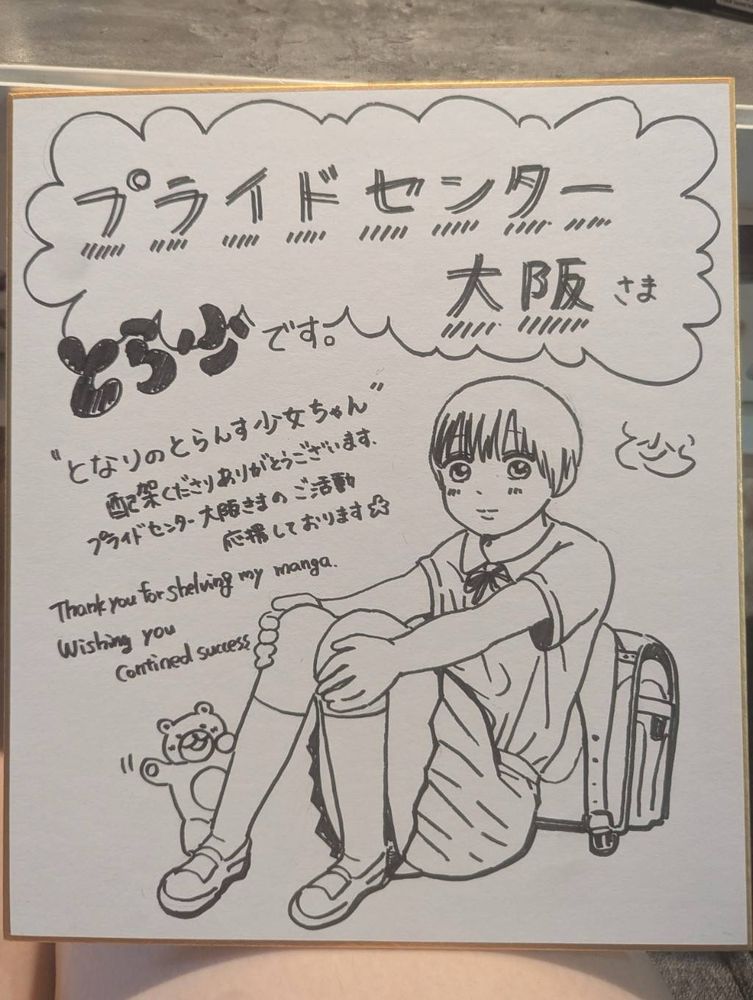 色紙に、とら少さんのイラストと以下のメッセージ書かせれている。

プライドセンター大阪さま
とら少です。
“となりのとらんす少女ちゃん”
配架くださりありがとうございます。
プライドセンター大阪きまのご活動
応援しております⭐︎
Thank you for shelving my manga.
Wishing you contined success.