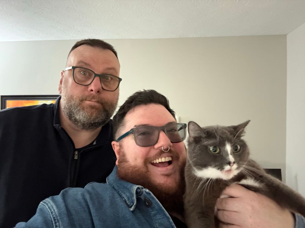 A very tall bearded man who looks confused standing behind a shorter bearded man who is very excited and holding a grey and white cat who visibly does not want to be involved in this selfie