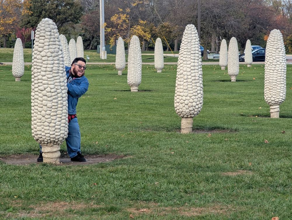 A fat beardy guy in sunglasses in a field of concrete corn cobs peaking from behind one of them with a big dumb smile 
