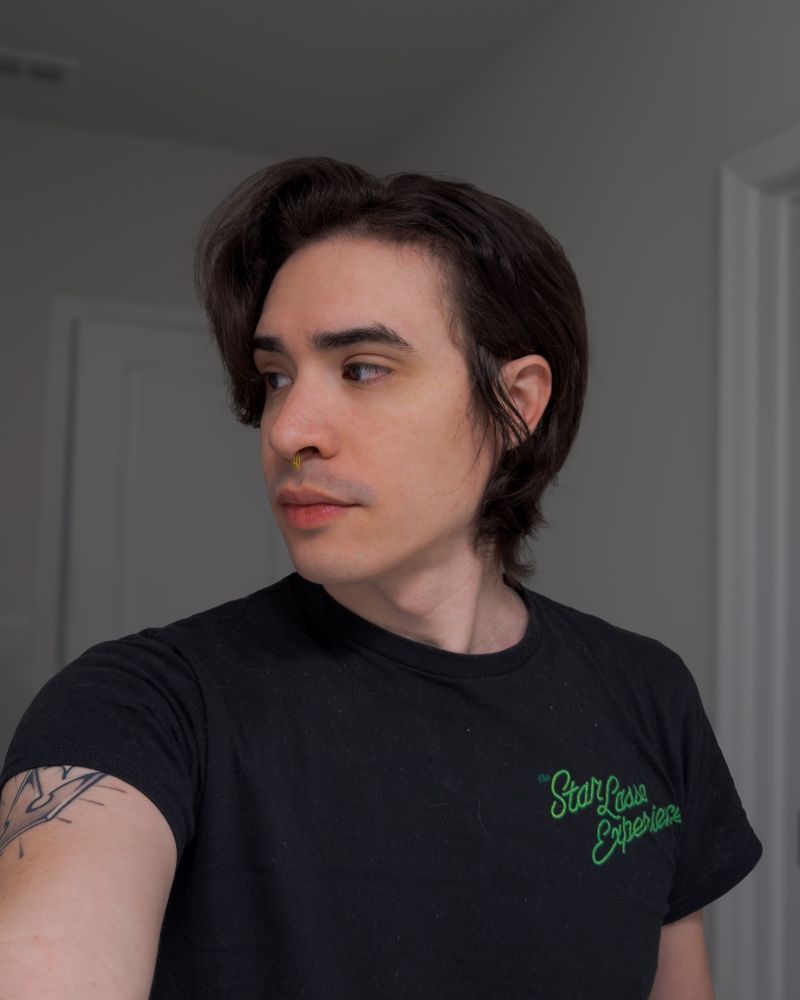 A selfie of me, looking off to the left of frame. I’m wearing a black shirt with green lettering over the heart that says The Star Lasso Experience, and my hair is actually cooperating for once by letting me tuck part of it behind my ear. Nature is healing (my hair is getting longer again)