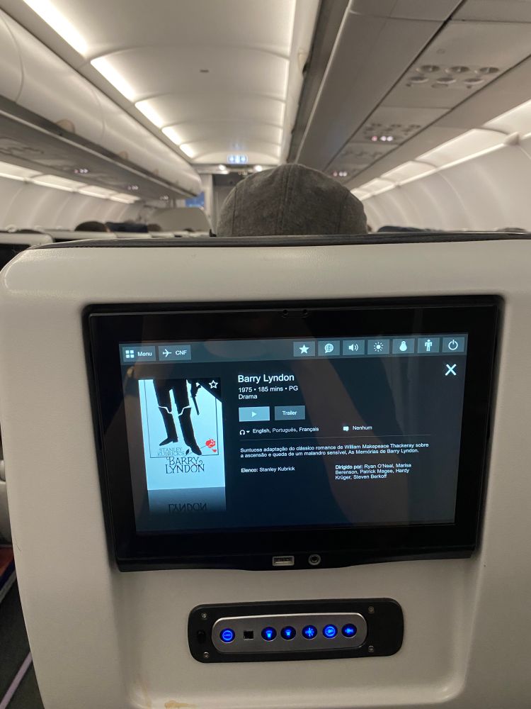 A cena é o ponto de vista de um passageiro de avião. Em destaque, um display digital de aproximadamente um palmo de tamanho, com as informações do filme “Barry Lyndon” no centro da tela acoplada no verso de um assento. Ao fundo, fora de foco, o interior cinzento do avião, iluminado por luzes frias.