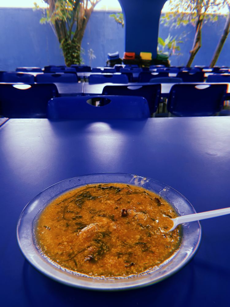 Um prato de vidro transparente servido com uma porção de canjica de milho salgada repousa sobre uma mesa azul num refeitório repleto de mesas e cadeiras azuis. Uma colher descartável branca está mergulhada na canjica com o cabo apoiado para fora do prato. Ao fundo, quatro lixeiras coloridas (azul, vermelha, amarela e verde) de coleta de lixo para reciclagem e mais longe, numa área externa após uma larga passagem arqueada, duas amoreiras.