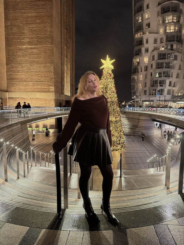 Photo of me! I’m standing at the top of some stairs outside in the evening, there’s a Christmas tree in the background. I’m wearing an off the shoulder red top, leather skirt and stiletto boots. 