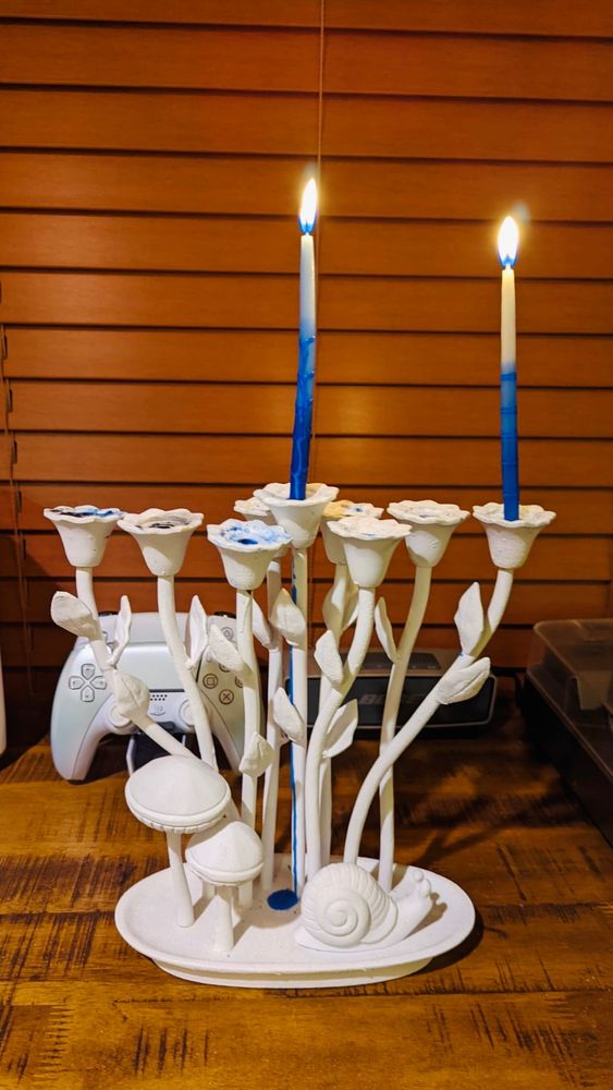 White menorah in the shape of a forest scene, with mushrooms, a snail, and tall flowers as the candle holders, in white, with the shamash and rightmost candles lit.