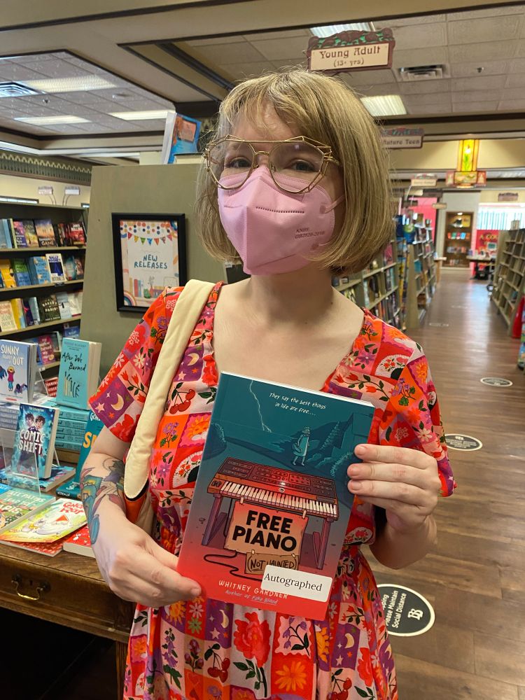 Whitney in a pink KN95 holding a newly autographed copy of Free Piano at Bolens Books