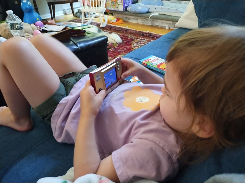 Toddler girl in lavender shirt with smiling flower on it reclines on a couch playing a Super Mario Bros Game & Watch