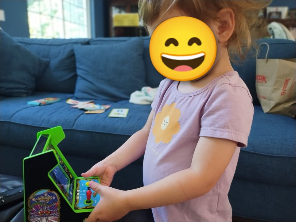 Toddler girl in lavender shirt with a smiling flower on it plays a mini Galaga arcade cabinet. Her face is obscured by a grinning emoji.