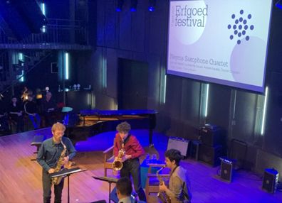 Saxophone quintet speelt op het podium met een groot scherm met Erfgoedfestival erachter.