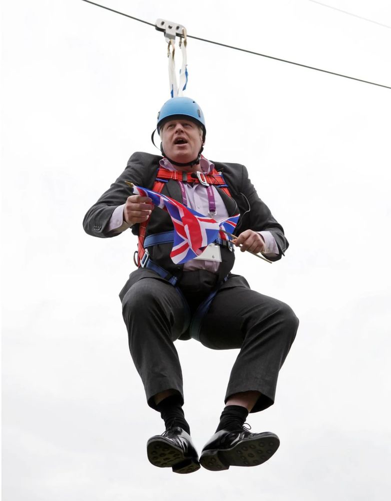 Boris Johnson hanging from a wire holding Union Jacks & wearing a safety helmet
