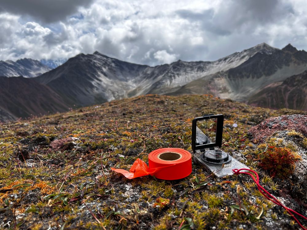 A roll of signal-orange flagging tape sits next to an open compass with reflecting mirror on the summit of an alpine peak in Yunnan, China which is covered in moss, orange lichen, and alpine vascular plants. In the backgrounds, roiling clouds and the distant peaks of the Hengduan.