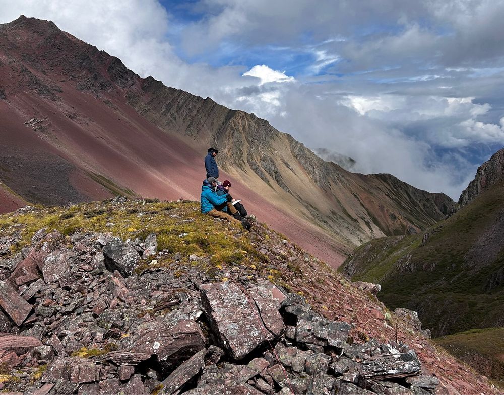 Three ecologists sitting on a rocky peak in SW China looking at field notes, mountain slopes and clouds in the background. Yunnan, China, 2024, collecting data as part of the Plant Ecology and Adaptation Knowledge Systems program at Missouri Botanical Garden.