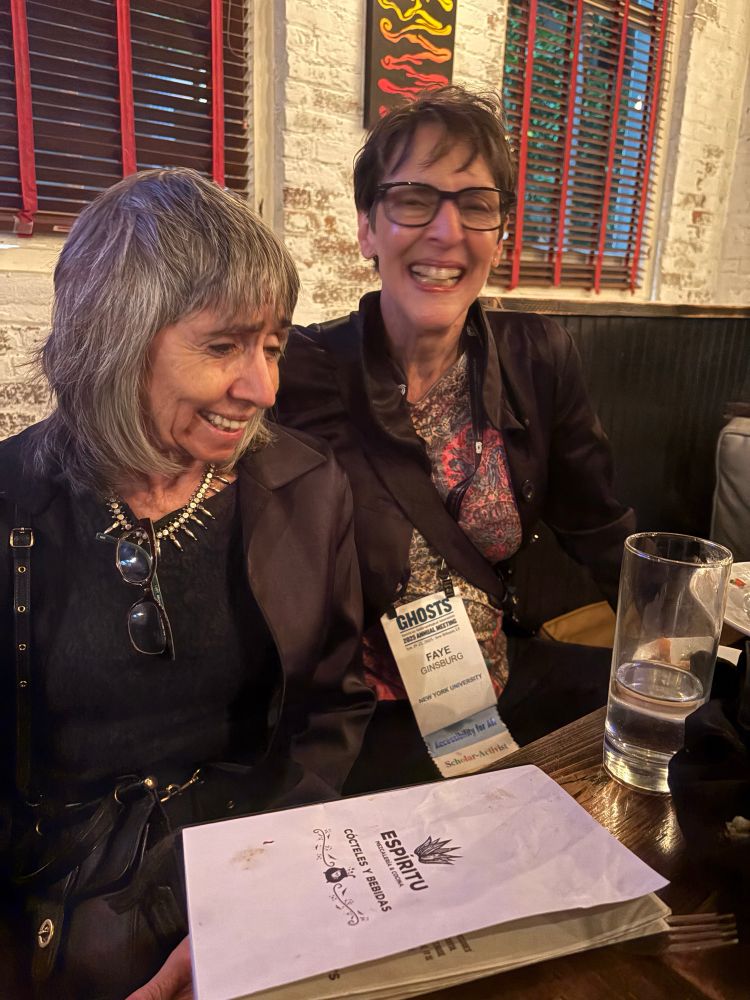 A white woman with shortish gray hair with a white woman with short hair and glasses sitting with a menu on the table. Both are smiling.