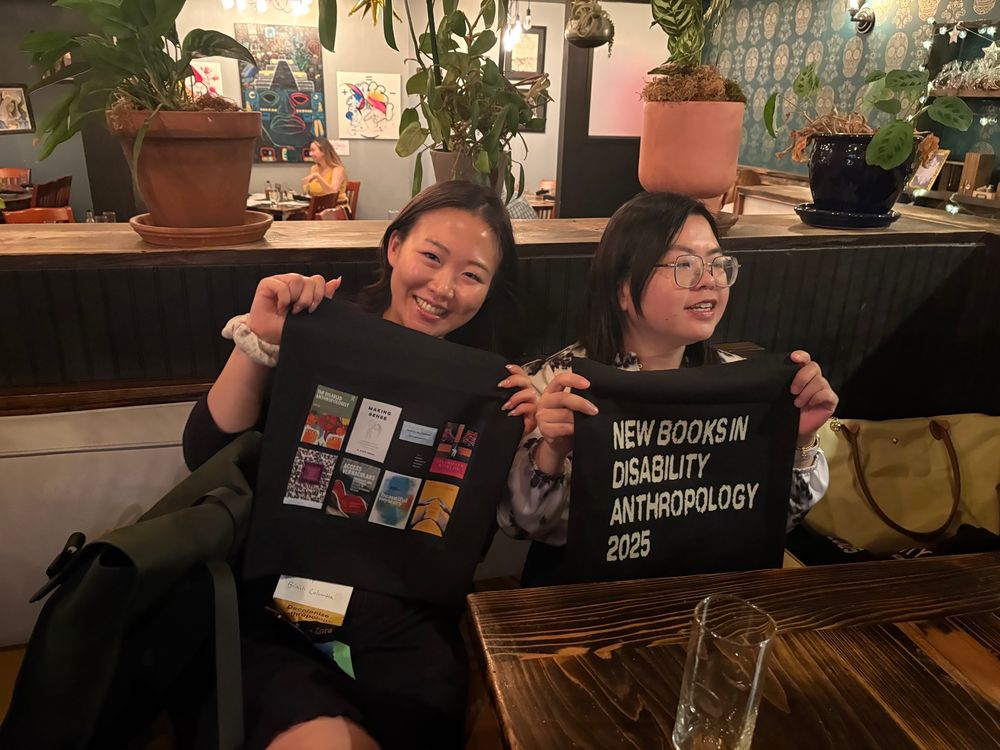Two asian women holding black tote bags — one with the front of the bag that reads “New Books in Disability Anthropology 2025” the other with the back of the same bag that has 8 different book covers printed on it