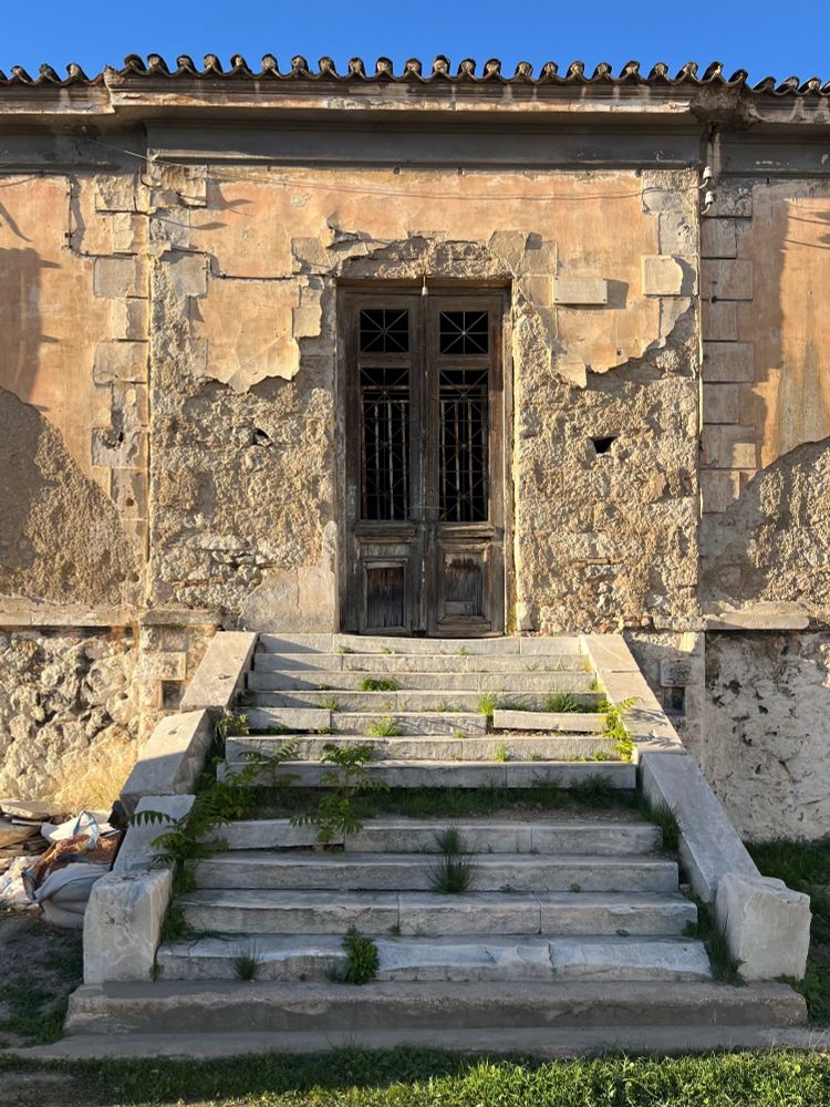 Colour photograph. In this larger neoclassical building, there’s a semi-basement work or storage space - such are often kitchens in structures of this period, and perhaps this was a mess hall. There are wide marble steps leading up to the front door, with ailanthus seedlings pushing their way through and dislodging the slabs. Up above, the plastered façade of the ground floor, still slightly tinted terracotta orange from the original mineral pigments, has mostly peeled off, showing the stone below. The wooden double door has inset panels of decorative grillework, now rusted, and with their accompanying glass gone. A wire and fitting for a long-gone lightbulb hangs at the top of the doorway.