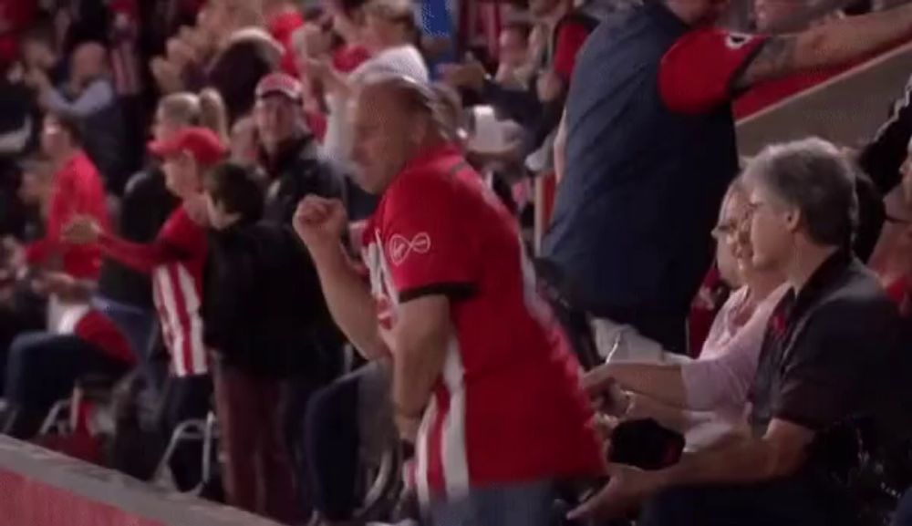 a man in a red shirt is jumping in the air while sitting in the stands at a basketball game .