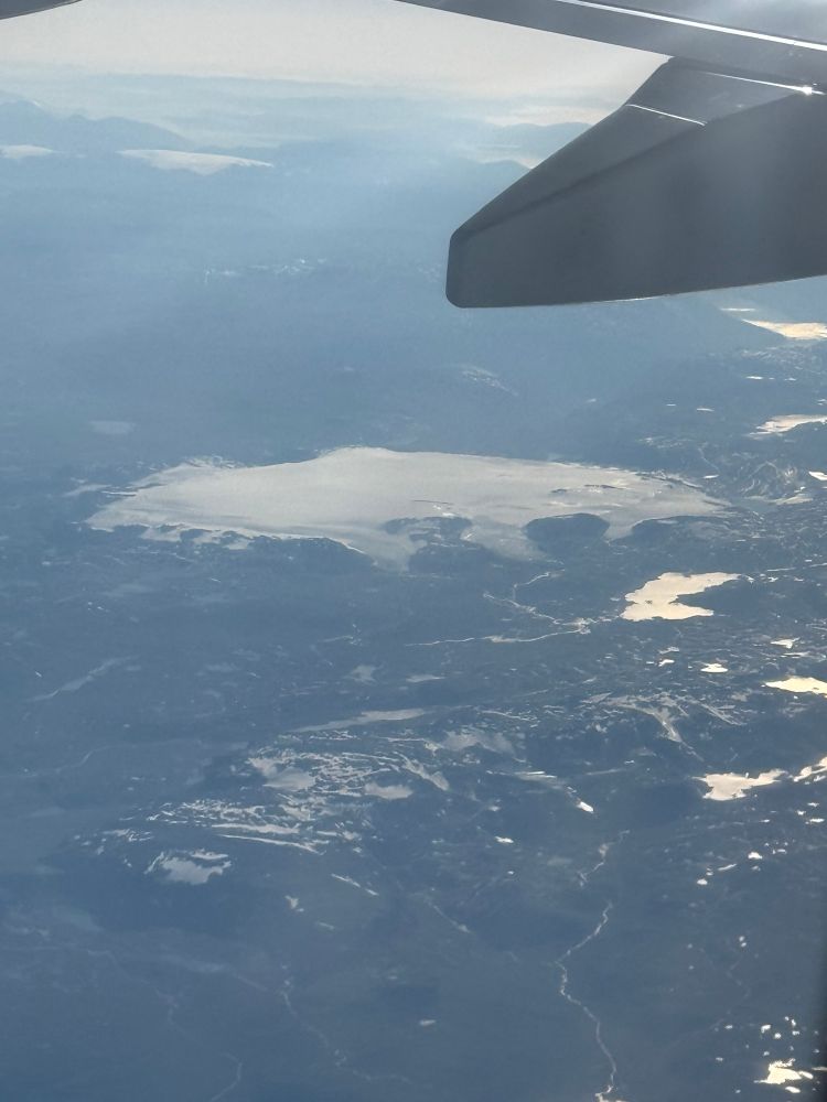An airplane window photo of an oval-shaped glacier in a mountainous setting. Several smaller tongues of ice extend downhill from the main glacier. There are some smaller patches of ice on the mountains in the foreground, and some liquid lakes shining brightly in the lower ground.