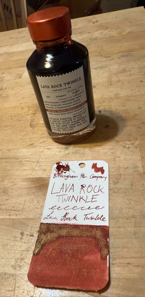 A swatch of dark red (with orangey-brown undertones) fountain pen ink, with gold shimmer on top of the red. The ink is from Birmingham Pen Company, and it is called Lava Rock Twinkle. The ink bottle is behind the swatch card. 
