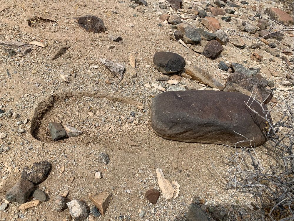 Some dark brown-gray rocks on a tan desert floor. In the center-right of the frame is a larger, semi-rectangular rock. To the left is an indentation in the ground shaped exactly the same way as that rock. There are no drag marks between the hole and the rock, suggesting that the rock was launched out of place. There were a lot of similarly displaced rocks around this area, which led to the question we addressed in the paper below: how did this happen?!