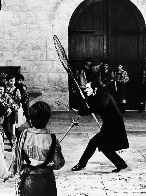 Photo of the Child-Catcher from Chitty Chitty Bang Bang.  Wearing long-ish black coat and holding up net.  Photo is from the side as he faces off against some of the children trying to escape.  