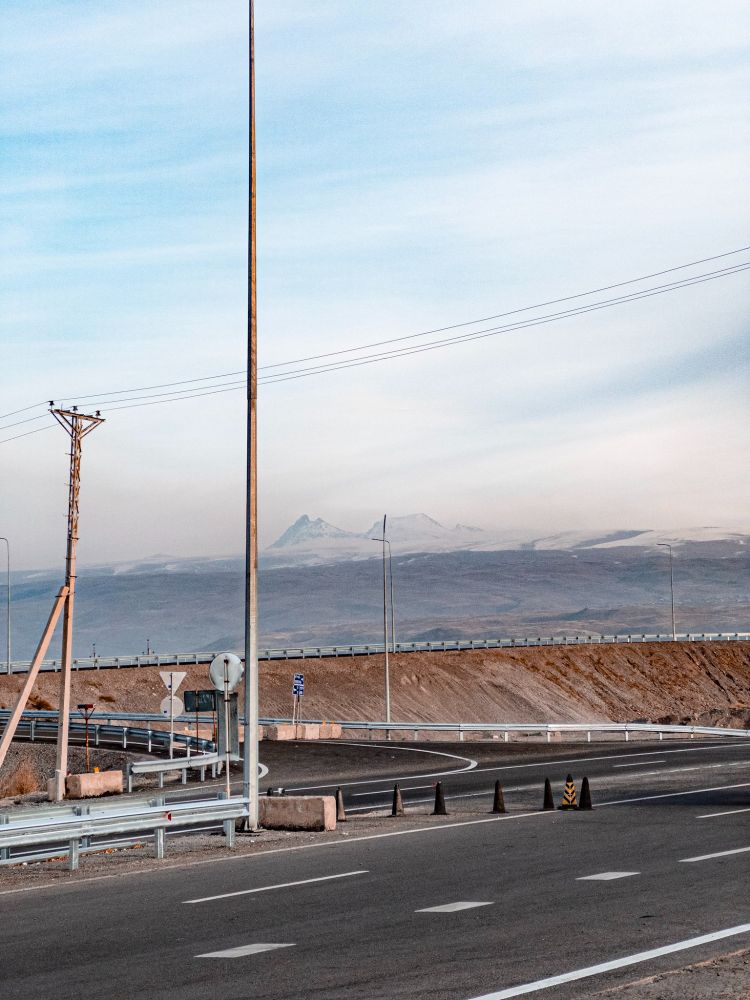 View from beside a highway, interchange crossing the frame horizontally, framing a hazy Mount Aragats in the distance