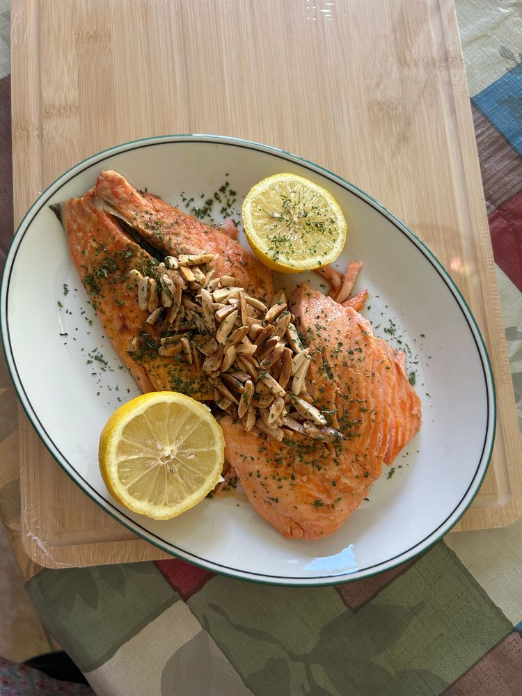 Pink trout fillet with slivered almonds in browned butter sprinkled with parsley and lemon garnish on the side.